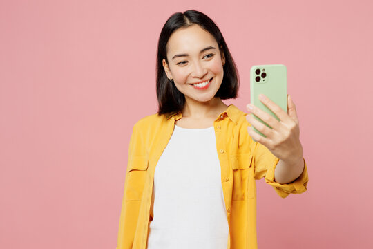 Young happy woman of Asian ethnicity wear yellow shirt white t-shirt doing selfie shot on mobile cell phone post photo on social network isolated on plain pastel light pink background studio portrait.