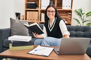 Young beautiful hispanic woman business worker using laptop reading book at office
