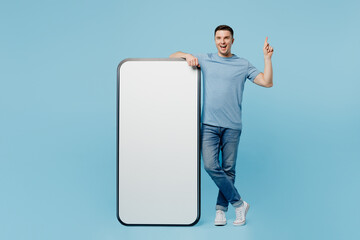 Full body young happy man wear casual t-shirt big huge blank screen mobile cell phone with workspace copy space mockup area point index finger up isolated on plain pastel light blue cyan background.