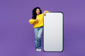 Full body happy young woman of African American ethnicity wear casual yellow sweater point finger on big huge blank screen mobile cell phone smartphone with area isolated on plain purple background.