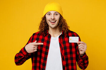 Young siling cool man wear red checkered shirt white t-shirt hat hold in hand point index finger on mock up of credit bank card isolated on plain yellow background studio portrait. Lifestyle concept.