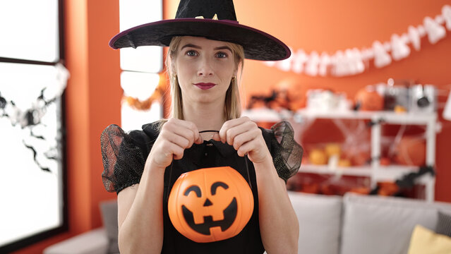 Young blonde woman wearing witch costume holding pumpkin basket at home