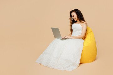 Full body happy young IT woman bride wear wedding dress posing sit in bag chair hold use work on laptop pc computer isolated on plain pastel beige background studio portrait. Ceremony party concept.
