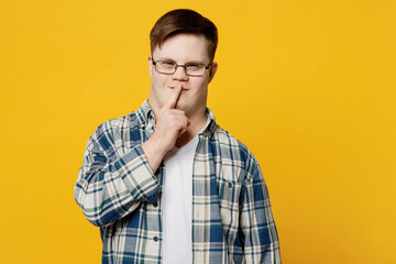 Young smiling man with down syndrome wear glasses casual clothes say hush be quiet with finger on lips shhh gesture isolated on pastel plain yellow color background. Genetic disease world day concept.