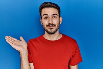 Young hispanic man with beard wearing red t shirt over blue background smiling cheerful presenting and pointing with palm of hand looking at the camera.