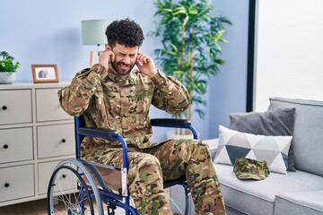 Fototapeta premium Arab man wearing camouflage army uniform sitting on wheelchair covering ears with fingers with annoyed expression for the noise of loud music. deaf concept.