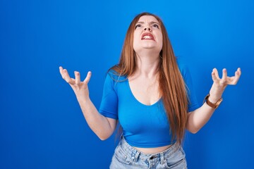 Redhead woman standing over blue background crazy and mad shouting and yelling with aggressive expression and arms raised. frustration concept.