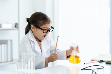 In chemistry classroom with many laboratory tools on shelves and table. A little Asian girl with glasses holding yellow chemical flask in her left hand look excited with new idea for next experiment.