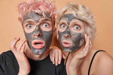 Impressed young woman and man apply facial clay mask for skin rejuvantaion feels scared of something keep mouth opened stand closely to each other isolated over brown background. Beauty procedures