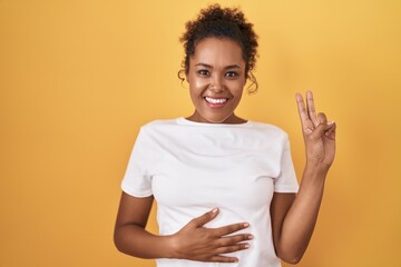 Young hispanic woman with curly hair standing over yellow background smiling swearing with hand on...