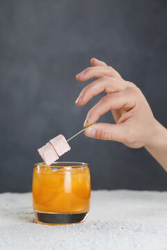 Bartender's hand is holding stick with marshmallow over orange cocktail at grey wall background.