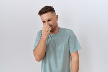 Hispanic man with beard standing over white background smelling something stinky and disgusting,...