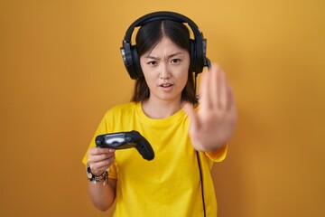 Chinese young woman playing video game holding controller doing stop gesture with hands palms, angry and frustration expression