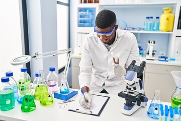 Young african american man wearing scientist uniform writing on clipboard holding test tubes at laboratory