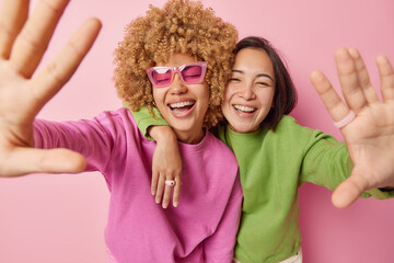 Positive overjoyed women keep arms outstreched laugh happily dressed in green and pink jumpers embrace and foolish around smile gladfully poses indoor. Friendship positive emotions and fun concept