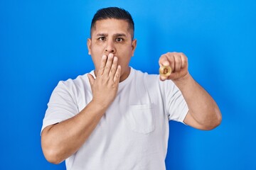 Hispanic young man holding virtual currency bitcoin covering mouth with hand, shocked and afraid...