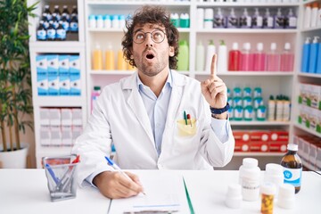 Hispanic young man working at pharmacy drugstore amazed and surprised looking up and pointing with fingers and raised arms.