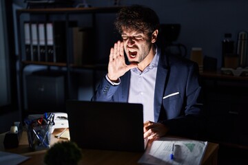 Hispanic young man working at the office at night shouting and screaming loud to side with hand on mouth. communication concept.