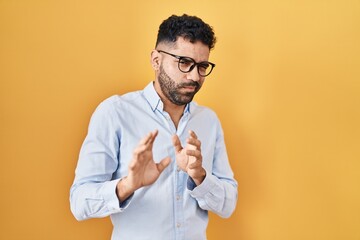 Hispanic man with beard standing over yellow background disgusted expression, displeased and fearful doing disgust face because aversion reaction. with hands raised
