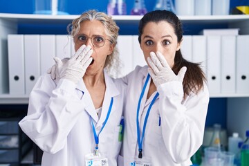 Mother and daughter working at scientist laboratory covering mouth with hand, shocked and afraid...