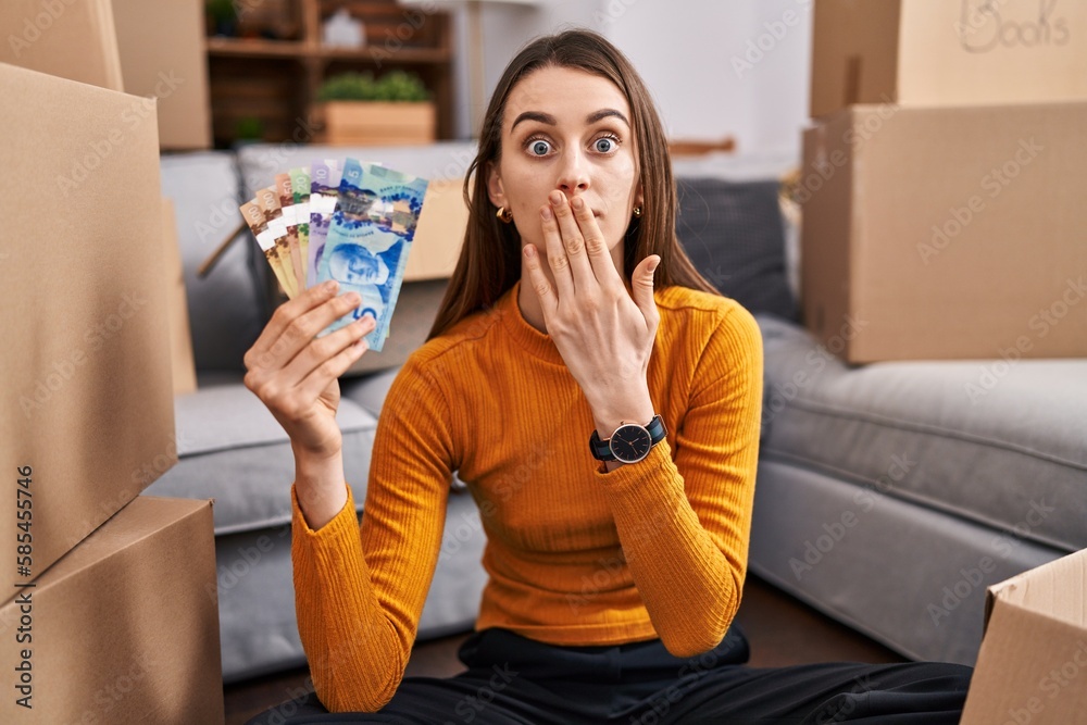 Poster Young caucasian woman sitting on the floor at new home holding canadian money covering mouth with hand, shocked and afraid for mistake. surprised expression