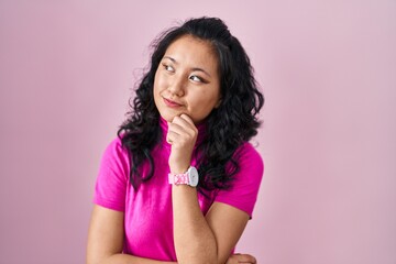 Young asian woman standing over pink background looking confident at the camera smiling with crossed arms and hand raised on chin. thinking positive.