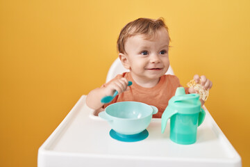 Adorable blond toddler sitting on highchair eating snack over isolated yellow background