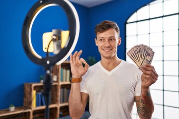 Caucasian man recording vlog tutorial with smartphone at home holding money doing ok sign with fingers, smiling friendly gesturing excellent symbol