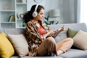 Curly-haired gir studentl sits on the sofa, wears headphones, looks in notebook and checks...