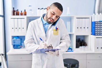 Young latin man scientist talking on smartphone writing document at laboratory