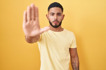 Young hispanic man standing over yellow background doing stop sing with palm of the hand. warning...