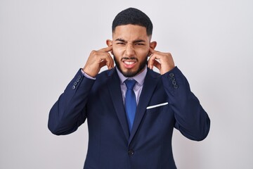 Young hispanic man wearing business suit and tie covering ears with fingers with annoyed expression for the noise of loud music. deaf concept.