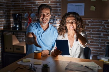 Middle age hispanic couple using touchpad sitting on the table at night smiling friendly offering handshake as greeting and welcoming. successful business.