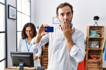 Hispanic man holding credit card at retail shop covering mouth with hand, shocked and afraid for...