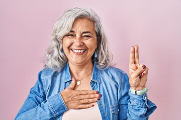 Middle age woman with grey hair standing over pink background smiling swearing with hand on chest...