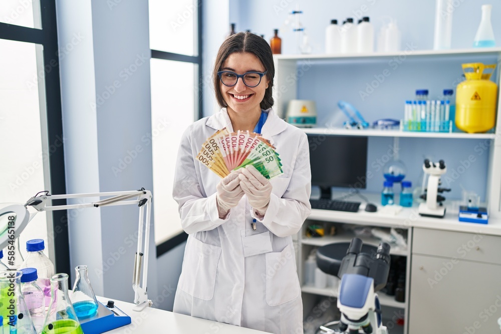 Poster Young hispanic woman working at scientist laboratory holding money banknotes winking looking at the camera with sexy expression, cheerful and happy face.