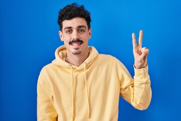 Naklejka premium Hispanic man standing over blue background showing and pointing up with fingers number two while smiling confident and happy.