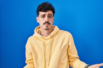 Obraz premium Hispanic man standing over blue background clueless and confused with open arms, no idea concept.