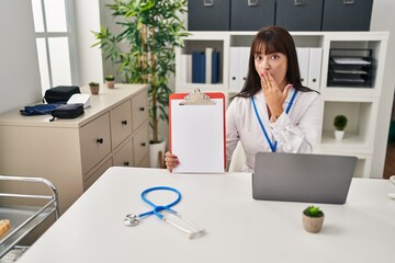 Young brunette doctor woman holding clipboard covering mouth with hand, shocked and afraid for...