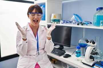 Young brunette woman working at scientist laboratory celebrating mad and crazy for success with arms raised and closed eyes screaming excited. winner concept