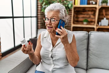 Muurstickers Senior grey-haired woman talking on the smartphone holding pills at home © Krakenimages.com