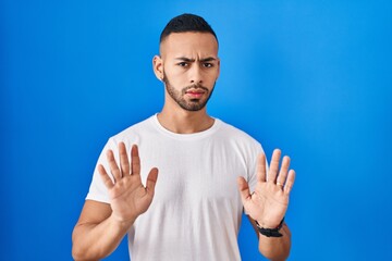 Young hispanic man standing over blue background moving away hands palms showing refusal and denial...
