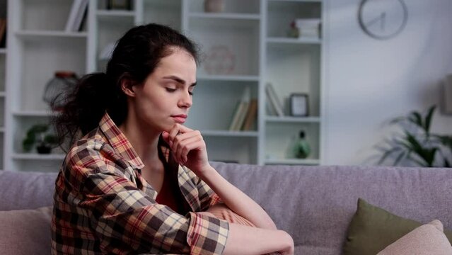 Young girl is deep in sad thoughts, thinks, looks away, feels tense and unhappy. Brunnete woman sitting on the couch have problen in personal life, feel depressed and broken.