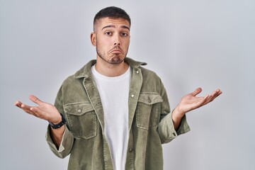 Young hispanic man standing over isolated background clueless and confused expression with arms and...