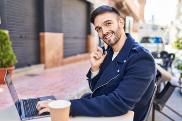 Young hispanic man talking on smartphone using laptop sitting on table at coffee shop terrace