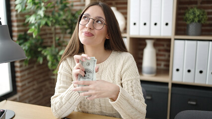 Young beautiful hispanic woman business worker holding dollars thinking at office