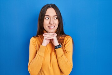 Young brunette woman standing over blue background laughing nervous and excited with hands on chin...
