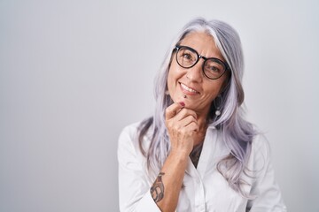 Middle age woman with tattoos wearing glasses standing over white background looking confident at the camera smiling with crossed arms and hand raised on chin. thinking positive.