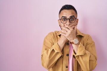 Young hispanic man standing over pink background shocked covering mouth with hands for mistake. secret concept.
