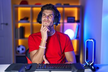 Young hispanic man playing video games with hand on chin thinking about question, pensive expression. smiling with thoughtful face. doubt concept.